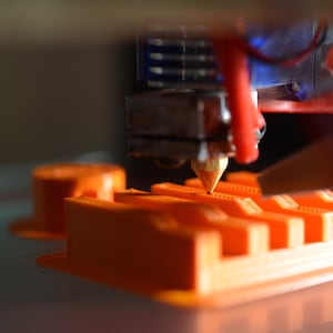 May include: Close-up of a 3D printer nozzle extruding orange plastic filament onto a platform, creating a layered, geometric design.