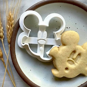 May include: White plastic cookie cutter shaped like Mickey Mouse with a Mickey Mouse shaped cookie on a brown speckled plate.