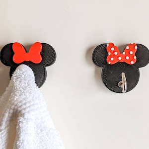 May include: Two black 3D printed Mickey Mouse head towel hooks with red bows. One hook has a white polka dot bow and the other has a solid red bow. One hook has a towel hanging from it.