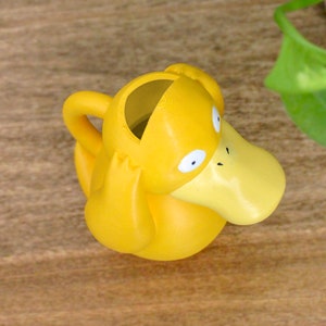May include: A yellow plastic watering can shaped like a duck with a wide, flat beak. The duck has large, round eyes and a small, black nose.