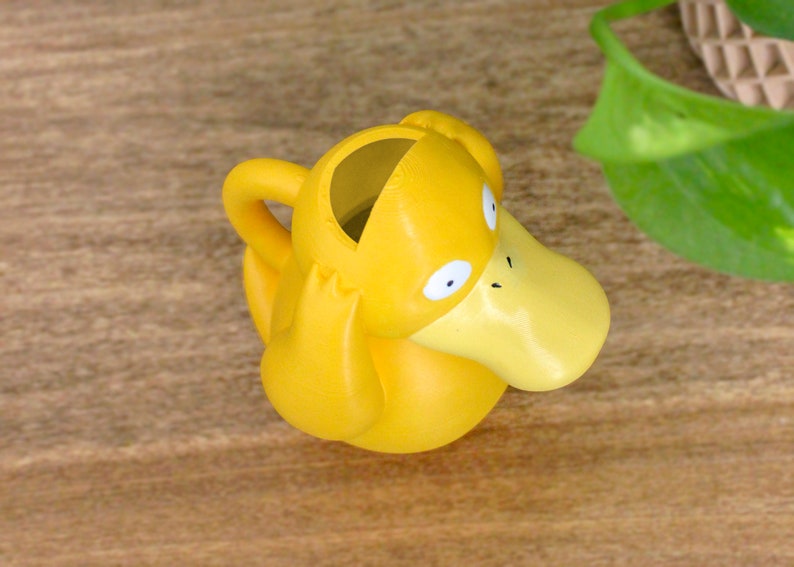 May include: A yellow plastic watering can shaped like a duck with a wide, flat beak. The duck has large, round eyes and a small, black nose.