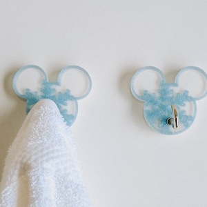 May include: Two light blue Mickey Mouse-shaped hooks with a snowflake design. One hook has a white towel hanging from it.