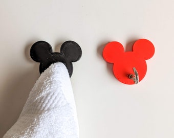 Op de afbeelding: Twee wandhaken in de vorm van Mickey Mouse oren. De ene haak is zwart en de andere is rood. Een witte handdoek hangt aan de zwarte haak.