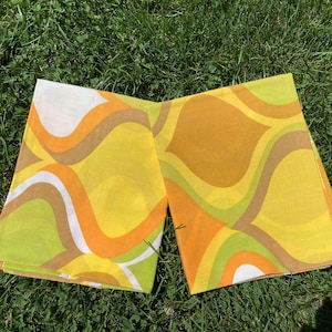 May include: Two folded vintage fabric napkins with a retro wavy pattern in yellow, orange, brown, white, and green. The napkins are lying on green grass.