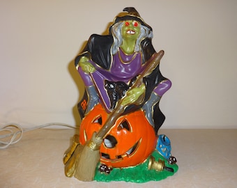 May include: A ceramic Halloween figurine of a witch sitting on a jack-o'-lantern. The witch is wearing a purple dress, a black cloak, and a pointed hat. She has green skin, white hair, and red eyes. She is holding a broom and a black cat is sitting on her lap.