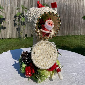 May include: A decorative Christmas mailbox made of white crocheted yarn, featuring a Santa Claus figurine inside. The mailbox is adorned with red and green accents, a red cardinal, and festive embellishments. The mailbox is sitting on a white surface.