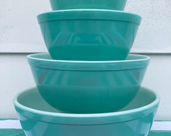 May include: A stack of four turquoise mixing bowls of varying sizes. The bowls are nested, with the largest bowl at the bottom and the smallest at the top. The bowls have a classic design with a rounded shape and a slight lip around the rim.