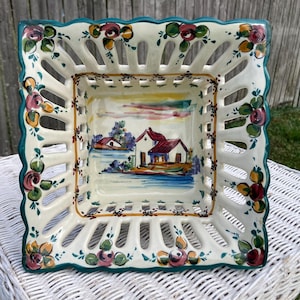 May include: Square ceramic basket with a scalloped edge, featuring a hand-painted landscape scene. The basket has a cream-colored base with a teal border and floral accents. The openwork design adds a decorative touch.