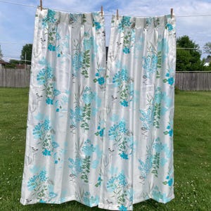 May include: Two white floral curtains with a blue and green botanical pattern hang from a clothesline. The curtains are made of a shiny fabric and are decorated with blue flowers and green leaves. The background is a green lawn and a cloudy sky.