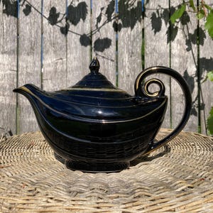May include: Black ceramic teapot with gold trim and a curved handle. The teapot has a lid with a finial and sits on a woven wicker surface. The background is a wooden fence with shadows of leaves.