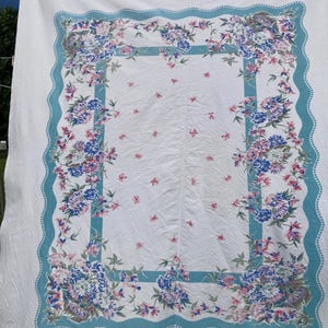 May include: White tablecloth with a floral design. The border features a teal scalloped edge and a teal rectangular frame with blue, pink, and purple flowers. Small pink flowers are scattered across the white center.