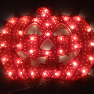 May include: A glowing, red plastic jack-o'-lantern decoration with a smiling face and star-shaped cutouts. The pumpkin is illuminated by small, warm white lights, creating a festive Halloween display. A power cord extends from the bottom.