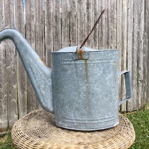 May include: A large, galvanized metal watering can with a long spout and a handle. The can has a rusty spout and a brown handle. It is sitting on a woven wicker surface, with a wooden fence in the background.