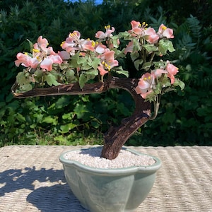 May include: A decorative bonsai tree sculpture featuring a dark brown textured trunk, light green leaves, and delicate pink flowers with yellow centers. The tree is set in a light green, scalloped ceramic pot filled with small white stones.