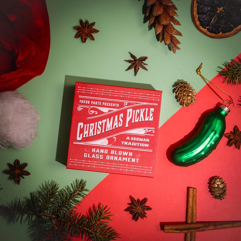 May include: Red box with white text that reads "Favor Parts Presents Christmas Pickle A German Tradition Hand Blown Glass Ornament". A green glass pickle ornament is on a red surface with cinnamon sticks, pine cones, and star anise.