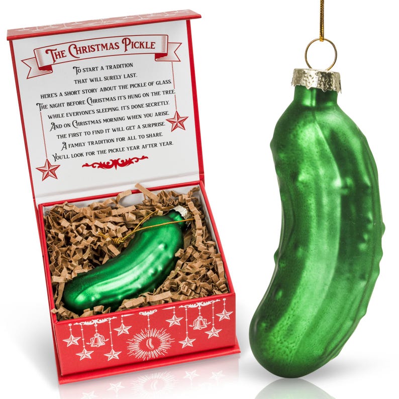May include: A green glass pickle ornament in a red box with a white background. The box has a gold ribbon and the words "The Christmas Pickle" on the front. The text inside the box explains the tradition of hiding a pickle ornament on the Christmas tree.