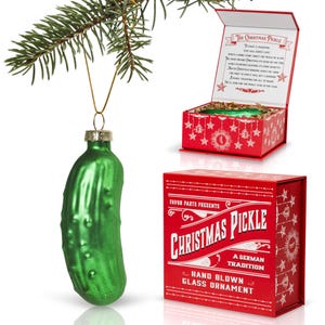 May include: A green glass pickle ornament with a red and white box that reads "Favor Parts Presents Christmas Pickle A German Tradition Hand Blown Glass Ornament".