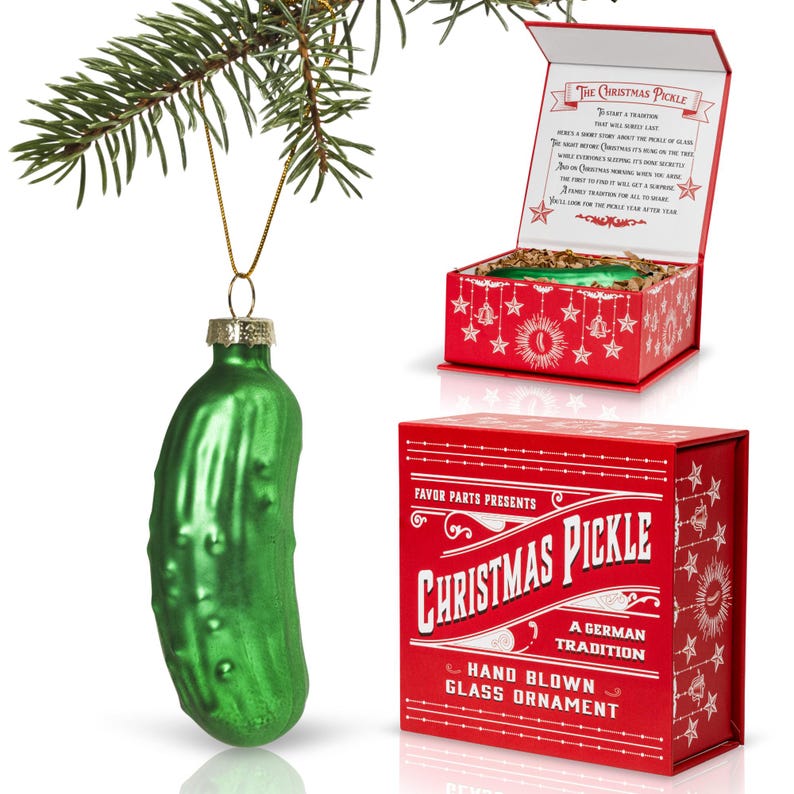 May include: A green glass pickle ornament with a red and white box that reads "Favor Parts Presents Christmas Pickle A German Tradition Hand Blown Glass Ornament".