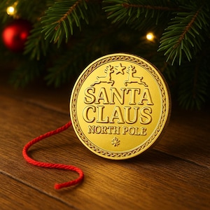 Könnte beinhalten: Eine goldfarbene Santa Claus-Münze mit dem Text "SANTA CLAUS NORTH POLE" und Rentier- und Sterndesigns. Ein roter Faden ist an der Münze befestigt. Die Münze liegt auf einer Holzoberfläche mit einem Weihnachtsbaum im Hintergrund.