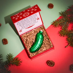 May include: A red box containing a Christmas pickle ornament. The box has a white background with a red border and a Christmas story printed on the front. The pickle ornament is green and has a shiny finish.