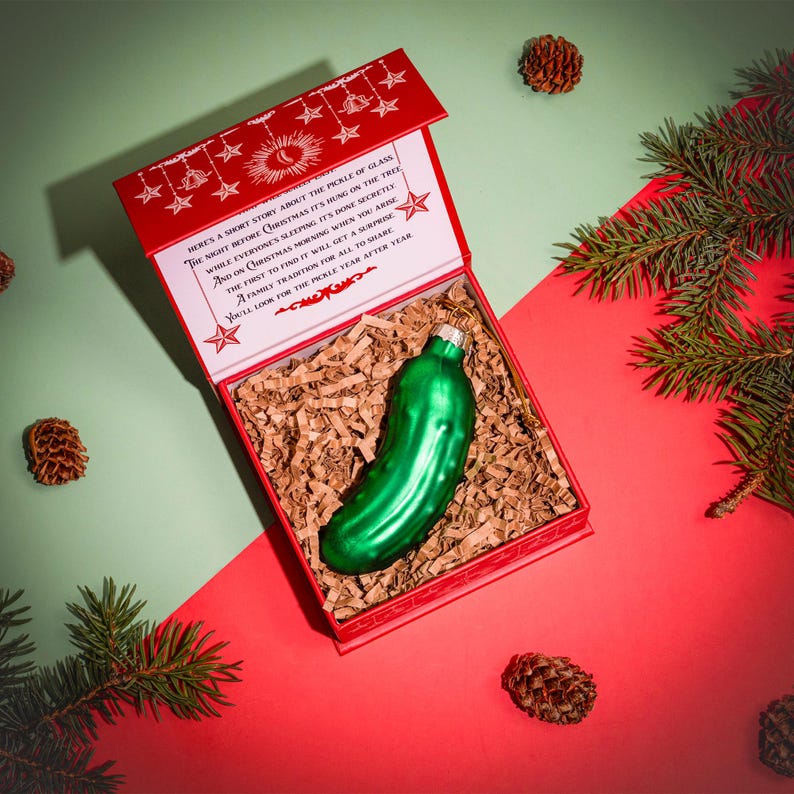 May include: A red box containing a Christmas pickle ornament. The box has a white background with a red border and a Christmas story printed on the front. The pickle ornament is green and has a shiny finish.