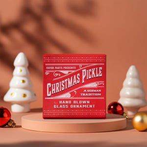 May include: Red box with white text that reads "Favor Parts Presents Christmas Pickle A German Tradition Hand Blown Glass Ornament". The box is sitting on a light brown surface with white ceramic Christmas trees and gold and red baubles.