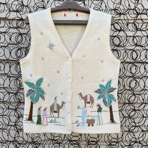 May include: A cream-colored knitted vest with a Christmas nativity scene embroidered on the front. The scene includes palm trees, three wise men, a camel, a star, and the baby Jesus in a manger.
