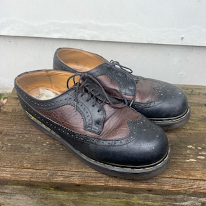 Può includere: Un paio di scarpe brogue in pelle nera e marrone con dettagli a punta ad ala. Le scarpe hanno una chiusura con lacci e una suola spessa in gomma nera.