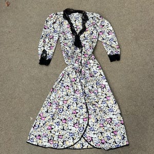 May include: A white floral print wrap dress with a black lace trim around the neckline and sleeves. The dress has a long, flowing skirt and a fitted bodice.