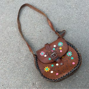 May include: A brown leather shoulder bag with a flap closure and a buckle. The bag is decorated with colorful flowers and white dots.