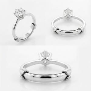 May include: A silver ring with a large, clear gemstone. The ring has a simple band with a decorative element on the sides. The ring is shown from multiple angles.