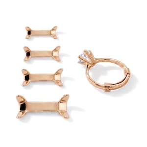 May include: A rose gold-toned ring with a clear, faceted stone in a prong setting, accompanied by four separate rose gold-toned ring size adjusters of varying lengths, each featuring a curved, bone-like shape designed to fit onto a ring band.