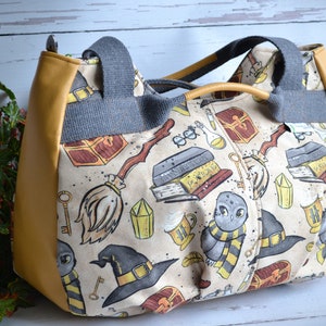 May include: A beige canvas tote bag with a yellow leather trim. The bag features a whimsical print of owls, broomsticks, keys, and other magical objects.