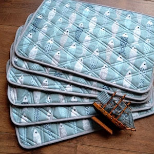 Puede incluir: Un juego de seis manteles individuales acolchados azul y blanco con un patrón de peces. Los manteles individuales tienen un borde gris y están apilados uno encima del otro.