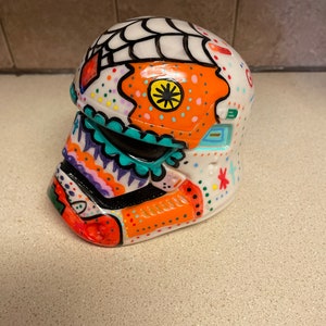 Op de afbeelding: Een witte, decoratieve helm, beschilderd met kleurrijke, op de Dag van de Doden ge&iuml;nspireerde ontwerpen. De helm heeft oranje, turquoise en paarse accenten, met een spinnenwebontwerp aan de bovenkant. Een uniek kunstwerk.