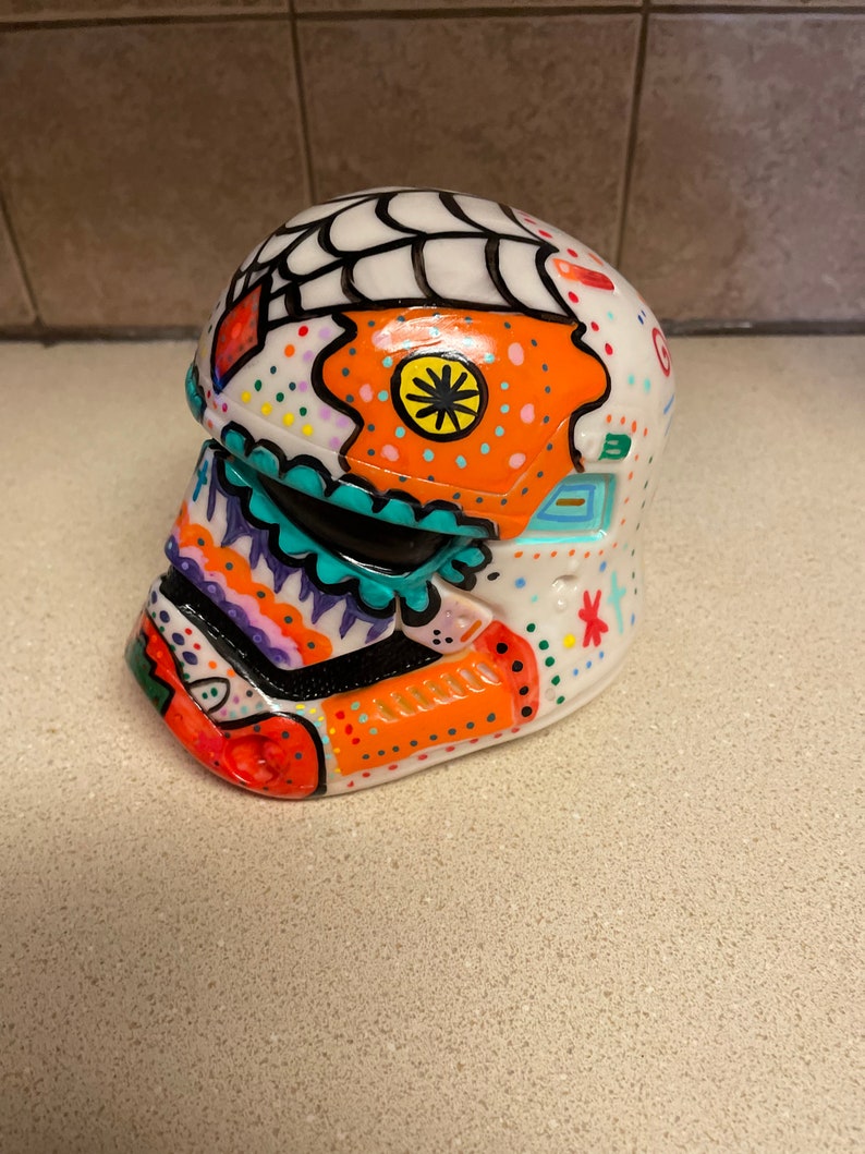 Op de afbeelding: Een witte, decoratieve helm, beschilderd met kleurrijke, op de Dag van de Doden ge&iuml;nspireerde ontwerpen. De helm heeft oranje, turquoise en paarse accenten, met een spinnenwebontwerp aan de bovenkant. Een uniek kunstwerk.