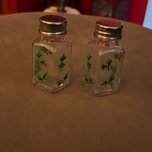 Op de afbeelding: Een paar zout- en peperstrooiers van helder glas met een vierkante voet en zilverkleurige metalen schroefdeksels. Elke strooier is voorzien van een handgeschilderd motief van groene hulstbladeren en kleine rode bessen op het glas.