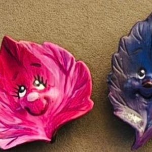 Op de afbeelding: Twee decoratieve bladvormige ornamenten met antropomorfe gezichten. Het ene blad is felroze met een glimlachende uitdrukking, terwijl het andere dieppaars is met een vergelijkbaar glimlachend gezicht. Beide items hebben gestructureerde oppervlakken.