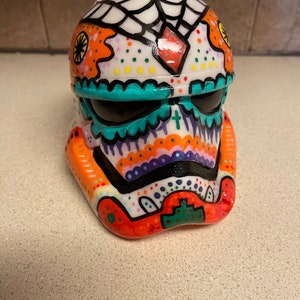 Op de afbeelding: Een decoratieve Stormtrooper-helm, met de hand beschilderd met een suikerschedelontwerp voor de Dag van de Doden. De helm heeft levendige kleuren, waaronder oranje, turquoise, paars en wit, met ingewikkelde patronen en een spinnenwebontwerp aan de bovenkant.