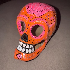 Op de afbeelding: Een handbeschilderd schedelbeeld met een feloranje basiskleur en levendige roze en zwarte stippenpatronen. De schedel heeft grote oogkassen, een zwarte neusholte en een kaaklijn met witte tanden die zijn omlijnd met zwarte inkt.