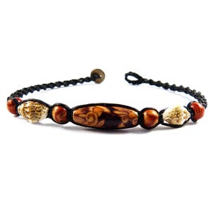 Könnte beinhalten: Ein Armband aus schwarzem Band mit braunen und weißen Muschelperlen und einer braunen Holzperle mit einem geschnitzten Design.