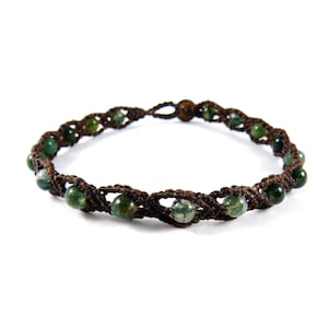 Puede incluir: Un brazalete trenzado marrón con cuentas de ágata verde. El brazalete está hecho con una técnica de anudado y tiene un diseño simple.