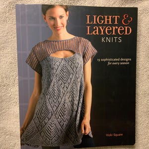 Pode incluir: Capa de livro com o título "Light & Layered Knits" em texto vermelho e branco. A capa apresenta uma mulher vestindo um top de malha cinza com um design de buraco de fechadura. O livro promete 19 designs sofisticados para cada estação.