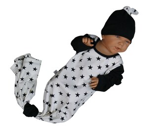 May include: A white and black baby sleep sack with a black star pattern. The sleep sack has a black knotted tail and the baby is wearing a black knitted hat.