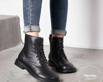 Op de afbeelding: Zwarte leren enkellaarsjes met vetersluiting en een zijrits. De laarzen hebben een ronde neus en een platte zool. Gedragen met een blauwe jeans. De merknaam "HerHis Handmade Shoes" is zichtbaar.