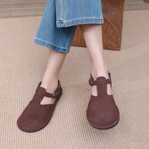 Peut inclure: Sandales en cuir marron avec une boucle et une bride ouverte. Les sandales sont portées par une personne portant un jean bleu.