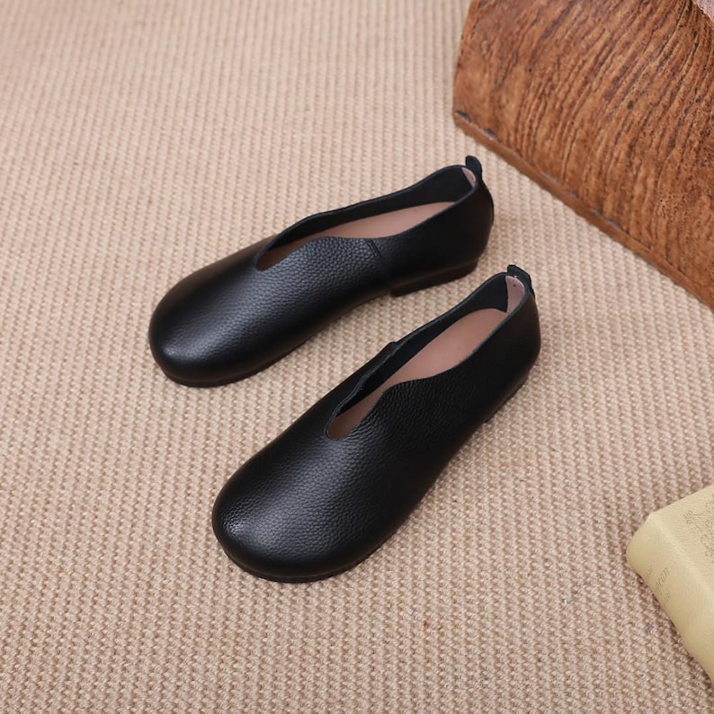 Op de afbeelding: Een paar zwarte leren ballerina's met een lage halslijn. De schoenen hebben een ronde neus en een kleine hak. Het leer heeft een textuur. De schoenen worden gepresenteerd op een neutraal gekleurd oppervlak.