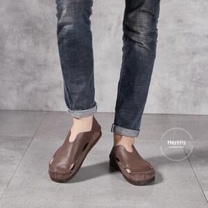 May include: Brown leather sandals with a cut-out design. The sandals have a closed toe and a strap across the top of the foot. The sandals are being worn by a person wearing blue jeans.