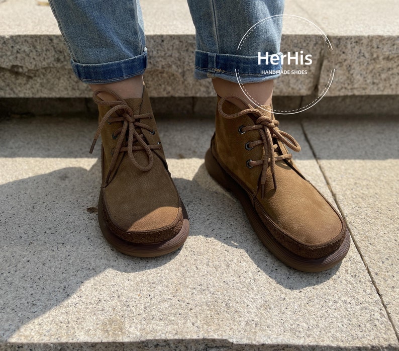 May include: Brown suede ankle boots with laces and a brown rubber sole. The boots are worn by a person wearing blue jeans. The text "HerHis HANDMADE SHOES" is visible on the image.