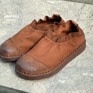 Op de afbeelding: Een paar bruin leren instappers. De schoenen hebben een ronde neus, een getextureerde wreef en een gerimpelde kraag. De zolen zijn donkerbruin met zichtbare stiksels. De schoenen worden gepresenteerd op een lichtgrijze ondergrond.